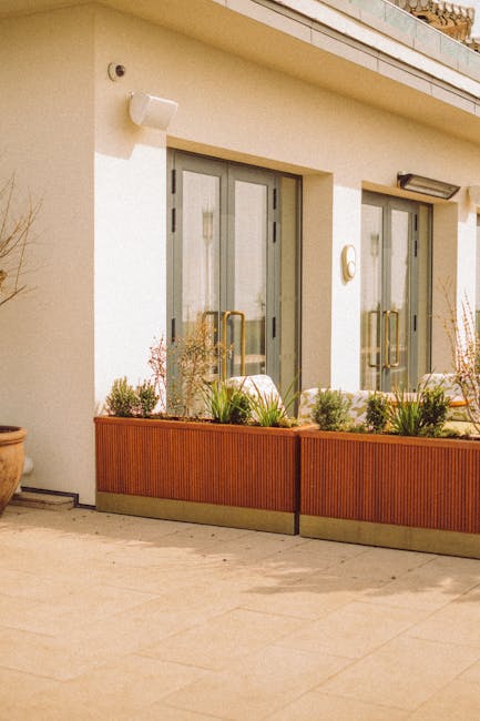 A detailed photograph of a rooftop terrace garden in Belgravia, showing lush green plants in wooden and stone planters, including a small hedge trimmed neatly along a pathway made of stone slabs. A pa